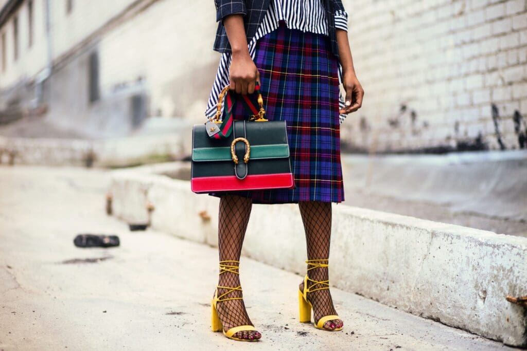 Picture of the legs and feet of a woman wearing yellow heels. She is also wearing a purse with read and green stripes.