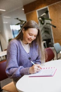 A woman wearing a purple sweater writing in her notebook.