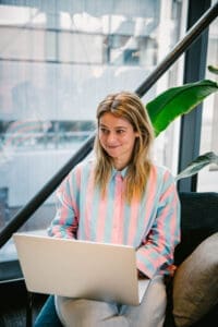 A woman with a pink and blue striped blouse writing on her laptop.